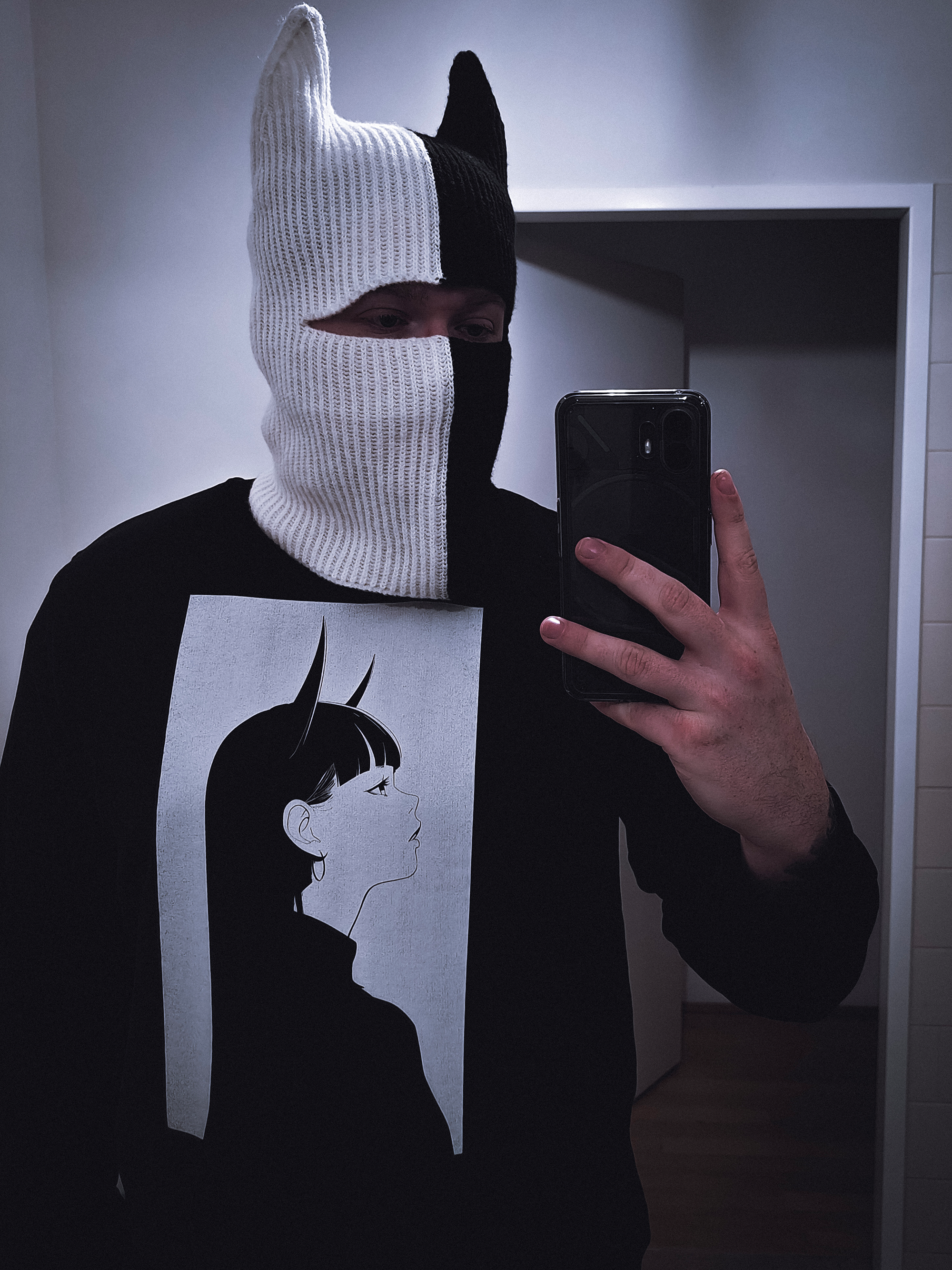 Person wearing a black sweatshirt featuring a minimalist anime design of a girl with long hair and horns, and a black and white knitted mask with pointed ears, taking a selfie in a mirror.