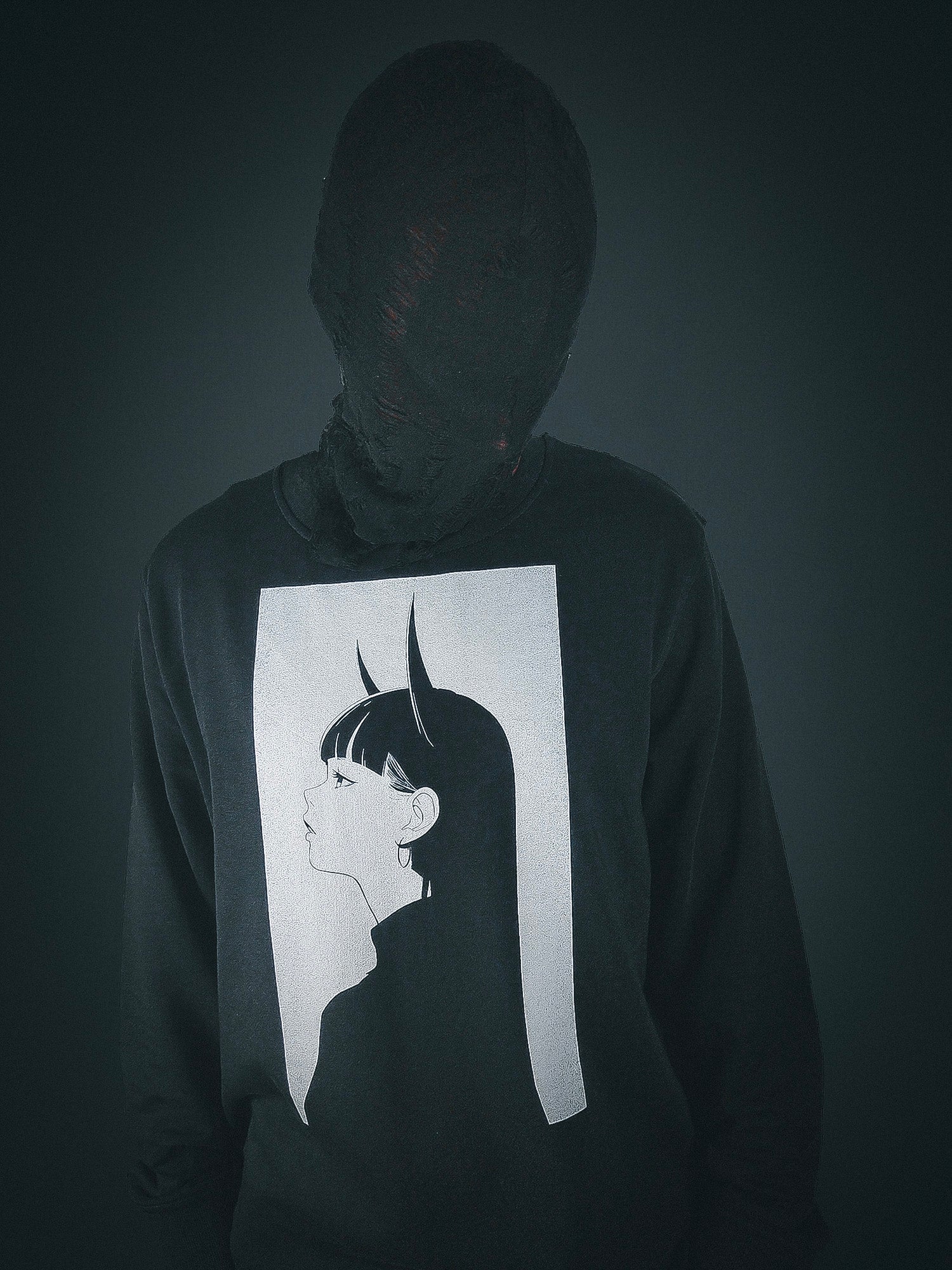 Person wearing a black sweatshirt featuring a minimalist anime design of a girl with long hair and horns, with their head wrapped in black fabric, standing against a dark background.